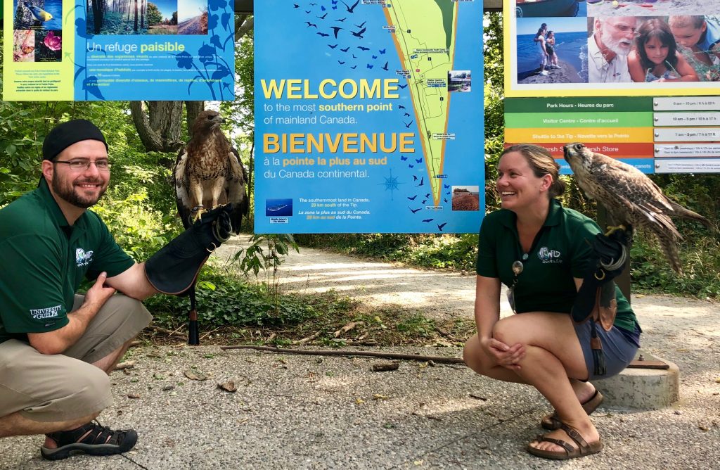 two handlers pose with birds by a park sign